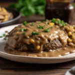 A plate of Amish Hamburger Steak Bake smothered in rich mushroom gravy, served with sautéed onions, corn, and fresh parsley on a rustic wooden table.